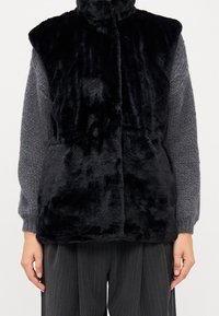 Gilet in pelliccia sintetica nera con colletto alto, chiusura frontale con bottoni e texture morbida. Abbinato a un maglione grigio lavorato a maglia e pantaloni a righe sottili.