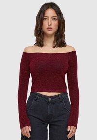Burgundi színű, off-shoulder crop top hosszú ujjakkal, fényes, texturált anyagból készült, magas derekú fekete farmernadrággal párosítva.