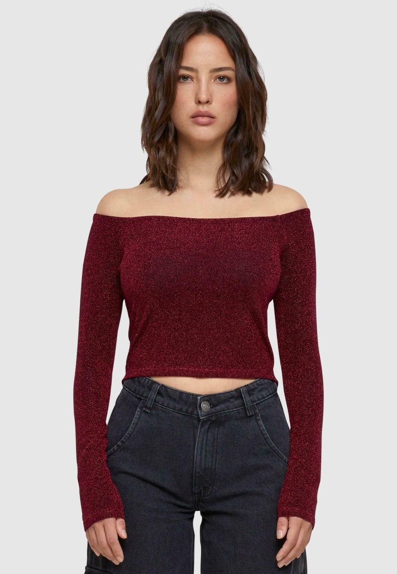 Burgundi színű, off-shoulder crop top hosszú ujjakkal, fényes, texturált anyagból készült, magas derekú fekete farmernadrággal párosítva.