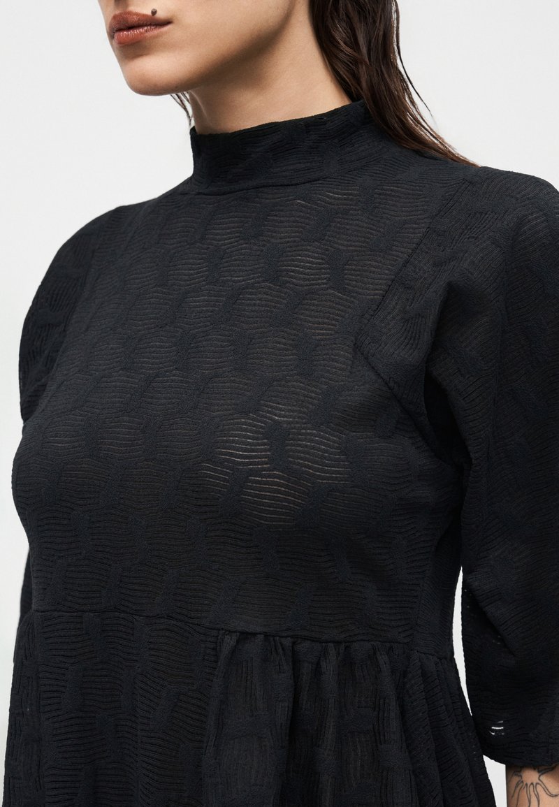 Frau trägt ein schwarzes, strukturiertes Kleid mit hohem Kragen, Puffärmeln und geraffter Taille, zeigt einen tätowierten Unterarm und Lippen mit einem Muttermal darüber.