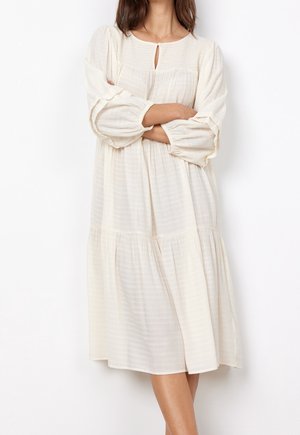 Femme portant une robe midi ample à manches longues couleur crème, avec un petit décolleté en forme de goutte et une taille froncée.