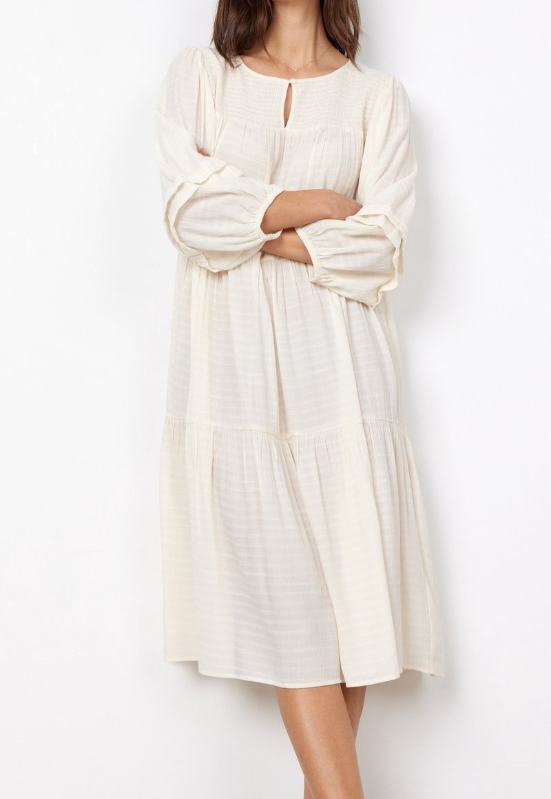 Femme portant une robe midi ample à manches longues couleur crème, avec un petit décolleté en forme de goutte et une taille froncée.