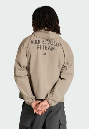 Person mit Dreadlocks, der eine beige Kapuzenjacke mit dem Aufdruck "AUDI REVOLUT F1 TEAM" auf dem Rücken trägt, die Hände hinter dem Rücken verschränkt, graue Hosen mit Taschen.