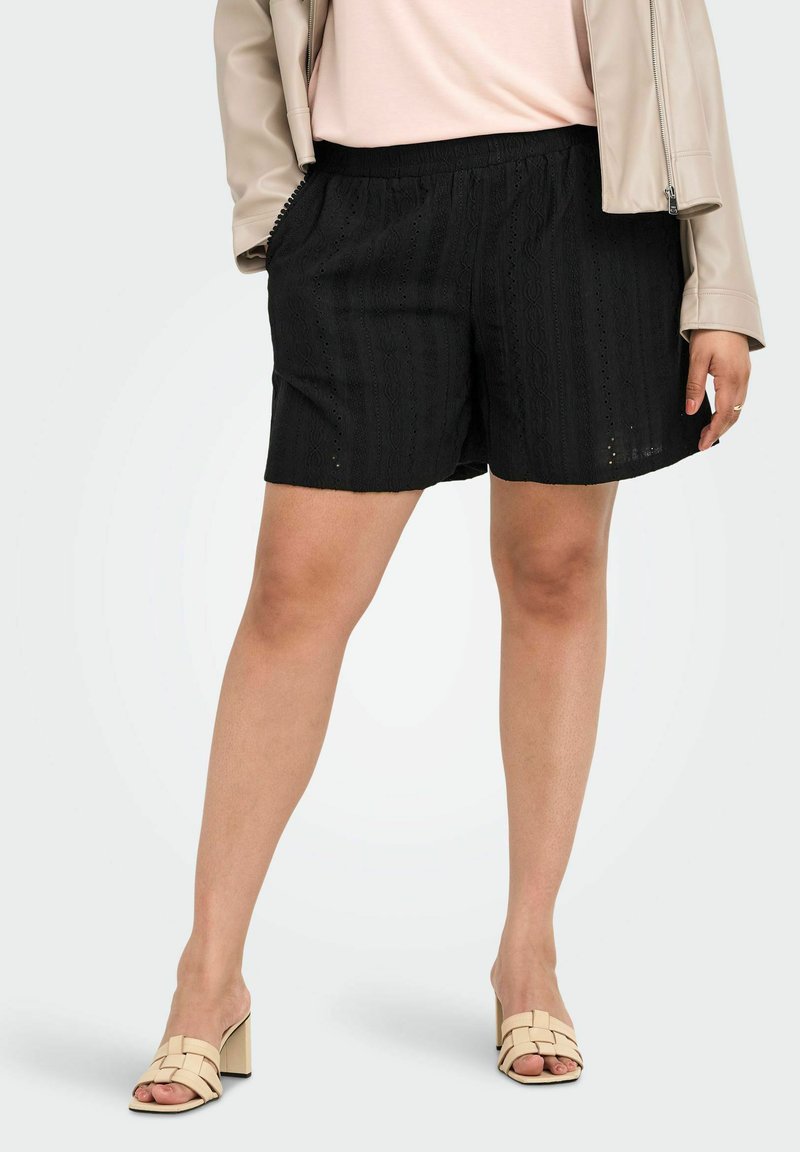 Femme portant un short noir texturé, des sandales beige à talons ouvertes, un haut rose pâle et une veste beige clair avec une main dans la poche.