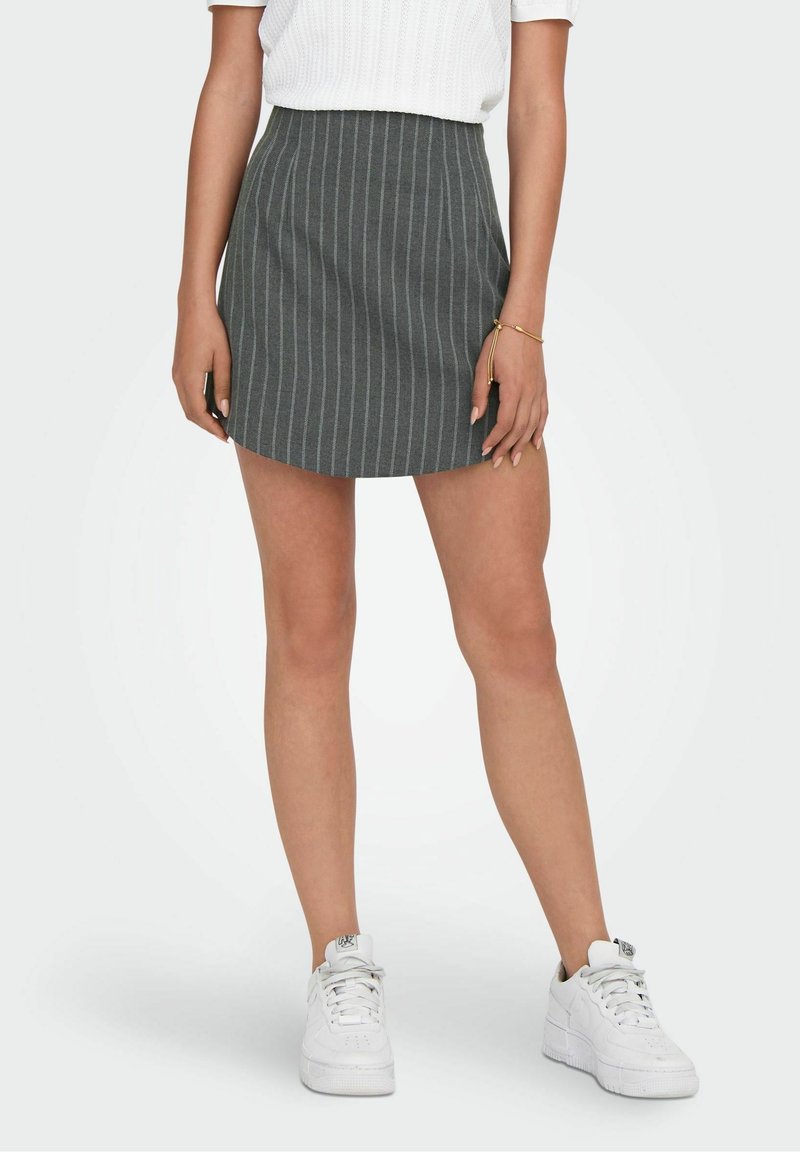 Femme portant une mini-jupe grise à fines rayures, un haut en maille blanc, des baskets blanches et un bracelet en or à son poignet gauche.