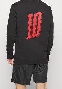 Sort sweatshirt med rødt tallet "10" grafik på ryggen. Stoffet virker blødt og har ribstrikkede manchetter og linning. Sorte shorts er synlige nedenunder.