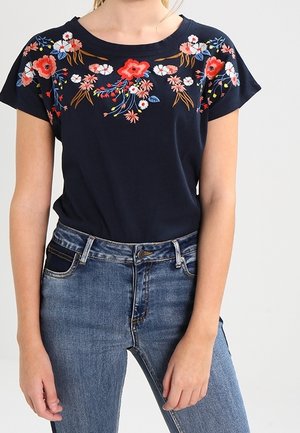 Granatowy top z krótkim rękawem ozdobiony żywym czerwonym, białym i różowym haftem kwiatowym na ramionach, zestawiony ze średnio niebieskimi dżinsami.