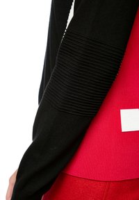 Schwarzer Strickpullover mit gerippten Details an den Ärmeln, über einem roten, gerippten Oberteil getragen. Mit einem weißen Logo-Patch auf dem roten Stoff.