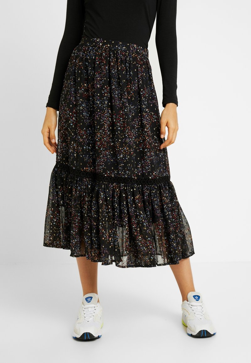 Jupe noire à motifs avec des détails floraux colorés, volants à l'ourlet, et en tissu léger et transparent. Associée à des baskets blanches.