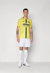 Kollane jalgpallimängija särk siniste detailidega ja "PAMESA cerámica" logoga, kombineeritud valgete pükste ja sokkidega, millel on tekstuuriga disain.