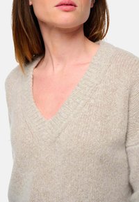 Hellbeige, weich strukturierter Pullover mit V-Ausschnitt und gerippten Kanten, der eine lockere Passform und subtile Farbvariationen aufweist.