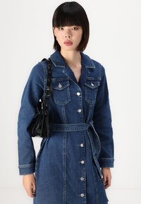 Τζάκετ καμπαρντίνα από τζιν σε σκούρο μπλε, με μακριά μανίκια, κουμπιά μπροστά, τσέπες στο στήθος και ζώνη στο ίδιο χρώμα. Διαθέτει ασημί αξεσουάρ και ραφή σε αντίθεση.