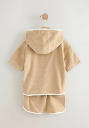 Completo da loungewear in terry cloth beige con maniche corte e pantaloni corti, dotato di cappuccio e piping bianco lungo i bordi. Texture morbida.