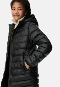 Cappotto invernale nero con cappuccio, caratterizzato da motivi trapuntati, tasche laterali e una cerniera frontale. Indossato sopra un maglione di colore chiaro.