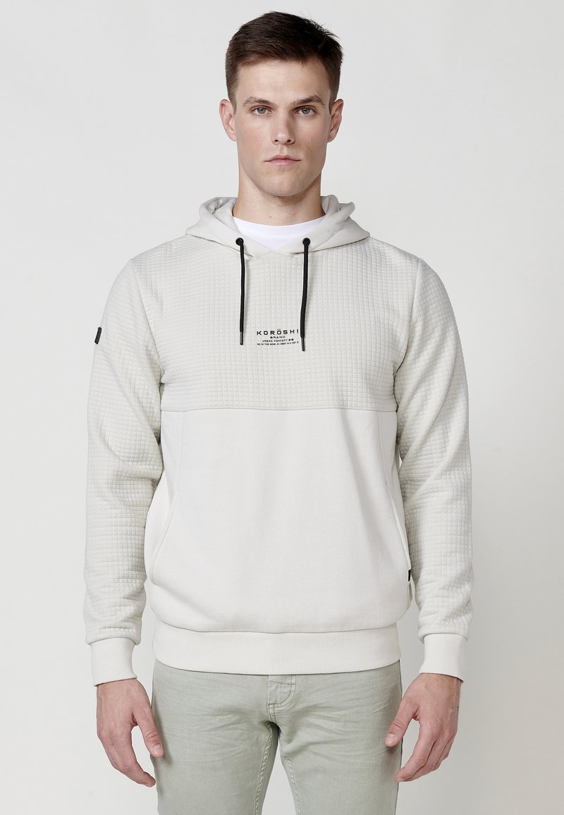 Hoodie grigio chiaro con un pannello superiore testurizzato, cappuccio standard, cordini neri e tasca frontale. Presenta il logo sul petto.