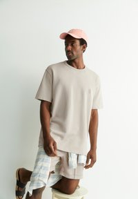 Lichtbeige katoenen t-shirt met een ronde hals, gedragen over lichte shorts met blauw-witte ruiten, gecombineerd met een roze pet en zwarte sandalen.
