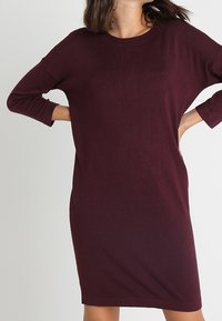 Vestido de punto burdeos con un corte relajado, cuello redondo y mangas de tres cuartos. Textura suave, largo hasta medio muslo y diseño sencillo.