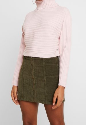 Persoon draagt een lichtroze geribbelde coltrui en een olijfgroene corduroy minirok met ritsdetail aan de voorkant.