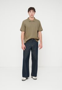 Πολύ κοντομάνικο πόλο σε ελιά πράσινο με ριμπ υφή, συνδυασμένο με σκούρο μπλε τζιν παντελόνι φαρδιάς γραμμής και λευκά αθλητικά παπούτσια. Απλός σχεδιασμός, casual εφαρμογή.