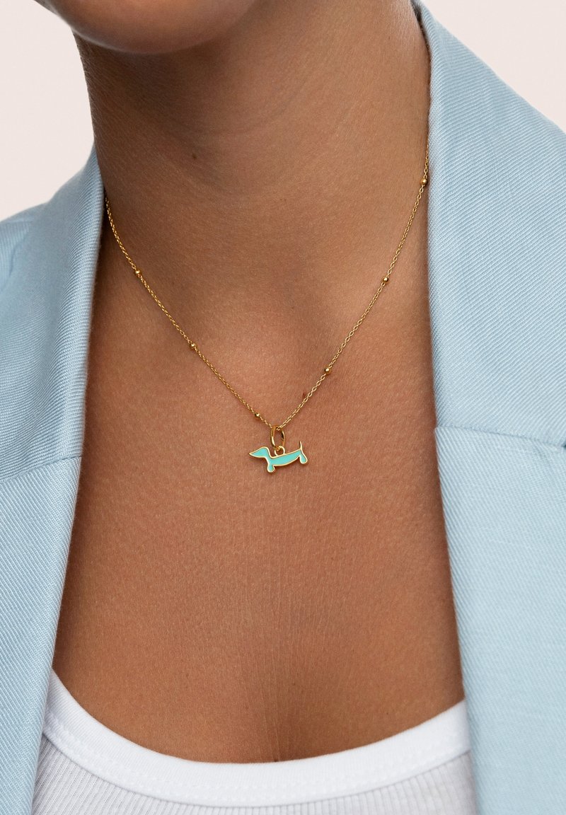 Collana di oro con piccole perle e un ciondolo a forma di bassotto in smalto turchese indossata sopra la pelle nuda e una maglietta bianca con un blazer azzurro chiaro.