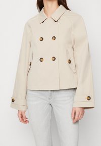 Chaqueta corta beige con diseño de doble botonadura, grandes botones marrones y mangas acampanadas. Fabricada en un tejido suave y estructurado.