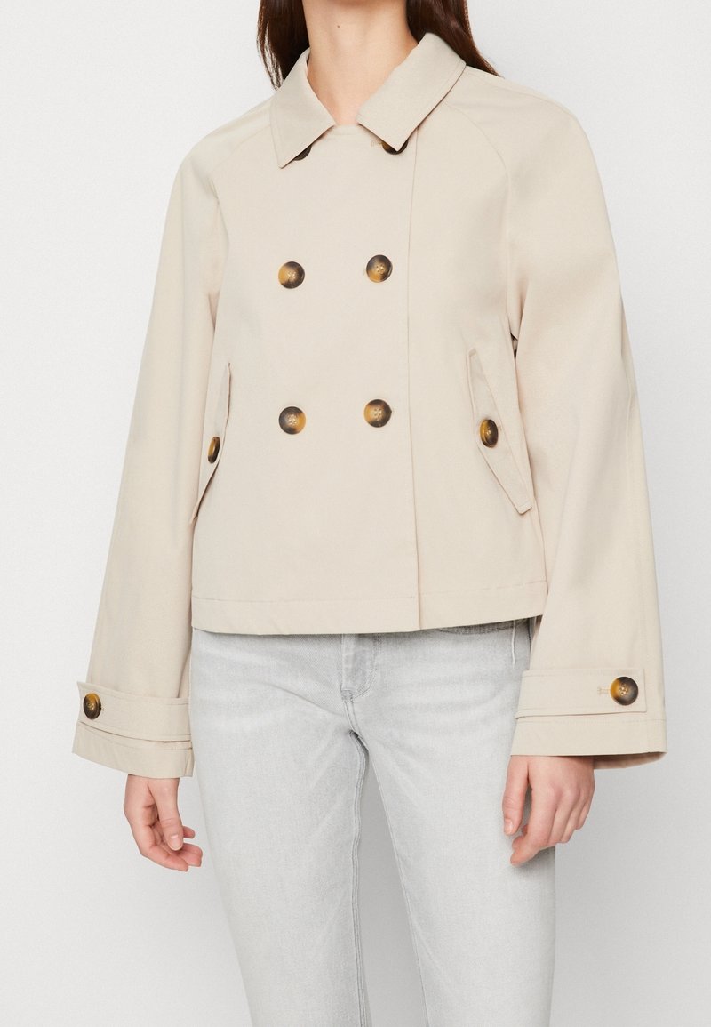 Chaqueta corta beige con diseño de doble botonadura, grandes botones marrones y mangas acampanadas. Fabricada en un tejido suave y estructurado.
