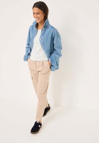 Jeansjacka i ljusblått med hög krage och knappstängning, kombinerad med en vit randig skjorta och beige cargobyxor. Svarta sneakers.