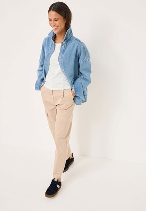 Giacca di jeans azzurro chiaro con colletto alto e chiusura a bottoni, abbinata a una camicia bianca a righe e pantaloni cargo beige. Sneakers nere.