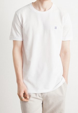 Mężczyzna ubrany w gładki biały t-shirt z krótkim rękawem z małym niebieskim "II" na lewej stronie klatki piersiowej, beżowe spodnie i pierścionek na lewej ręce.
