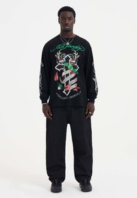 Zwarte longsleeve t-shirt met kleurrijk grafisch ontwerp, met tekst en motieven, gedragen met losse donkere broek en zwarte sneakers.
