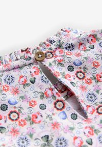 Nahaufnahme eines Stoffes mit kleinem roten, lila und grünem Blumenmuster, elastischem Rüschenkragen und einem einzelnen braunen Knopfverschluss.