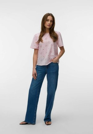 Vrouw staat en draagt een lichtroze blouse met korte mouwen en opengewerkte details, blauwe jeans met wijde pijpen en zwarte teenslippers tegen een effen witte achtergrond.