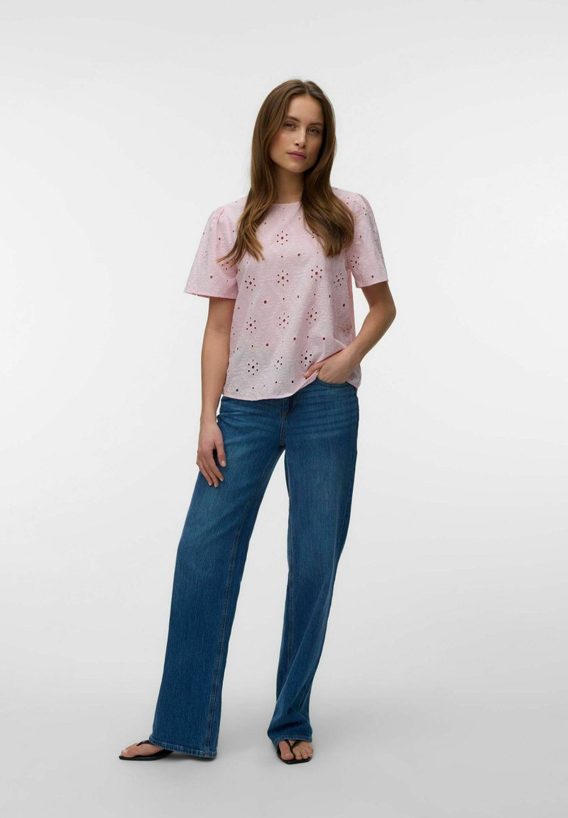 Femme debout portant un chemisier rose clair à manches courtes en broderie anglaise, un jean bleu à jambes larges et des tongs noires sur fond blanc uni.