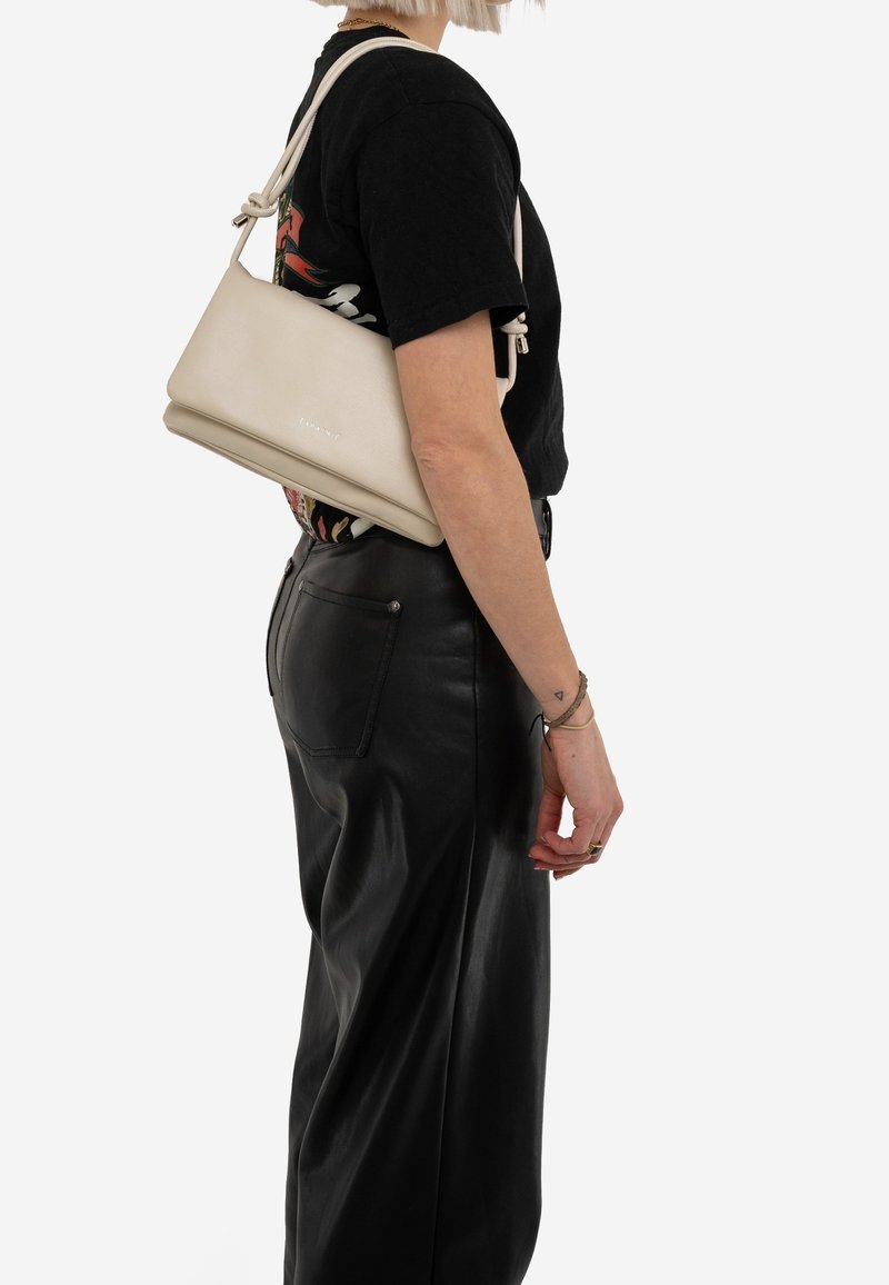 Personne portant un t-shirt noir et un pantalon en cuir noir, portant un sac à épaule beige avec une bandoulière nouée sur son épaule droite.