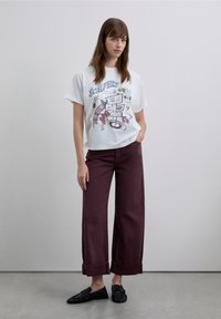 White t-shirt with graphic print and "SCALPERS" text, paired with wide burgundy trousers featuring rolled cuffs and black flat shoes.