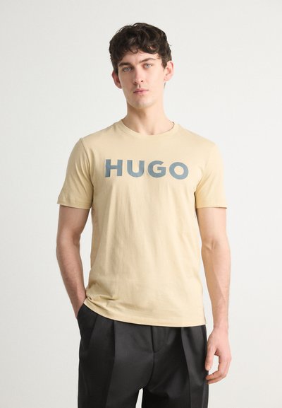 Бежова памучна тениска с къси ръкави, с големи сини букви, пишещи "HUGO" отпред. Свободенfit с кръгло деколте.