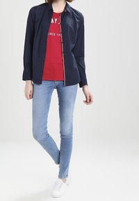 Granatowa koszula z długim rękawem na czerwonym koszulce z grafiką, zestawiona z jasnoniebieskimi rurkami i białymi trampkami.