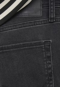 Primo piano della tasca posteriore di jeans in denim nero con dettaglio in tessuto a righe bianche e nere vicino al passante della cintura e etichetta cucita sopra la tasca.