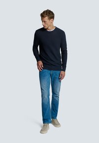 Marineblauwe gebreide trui met lange mouwen, gecombineerd met lichtblauwe spijkerbroek en beige schoenen. Model staat tegen een neutrale achtergrond.