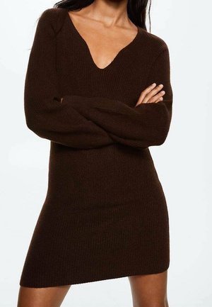 Femme portant une robe en maille marron foncé à manches longues avec un col en V, les bras croisés, sur un fond blanc uni.
