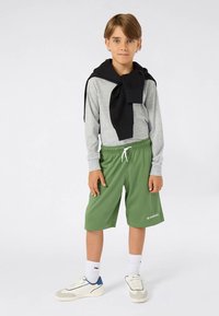 Chlapec stojící v šedém tričku s dlouhým rukávem, s černou mikinou uvázanou kolem ramen, v zelených šortkách, bílých ponožkách a bílých teniskách.