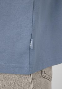 Ourlet d'une chemise en tissu bleu avec une petite étiquette "BZB" glissée dans la couture, reposant au-dessus de la poche d'un jean en denim gris clair.