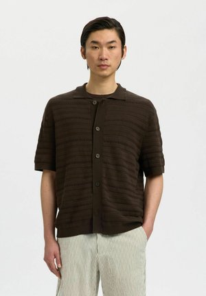 Mand iført en mørkebrun kortærmet knapbluse med strukturerede horisontale striber og lyse striped bukser, stående med den ene hånd i lommen.