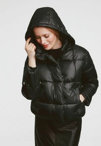 Kobieta w czarnej, ocieplanej kurtce z kapturem, trzymająca kaptur jedną ręką, a drugą w kieszeni, na jednolitym tle.
