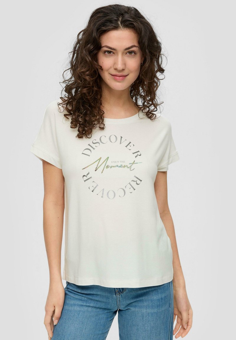 Helles Creme T-Shirt aus weichem Stoff, mit umgeschlagenen Ärmeln und Rundhalsausschnitt. Es hat ein kreisförmiges grafisches Textdesign in metallischen Farben.