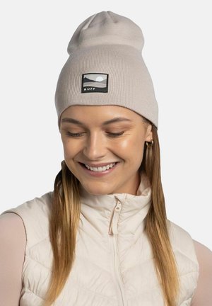 Beige knit beanie with a soft texture, featuring a black rectangular patch displaying a mountain and sun design, worn with a light vest.