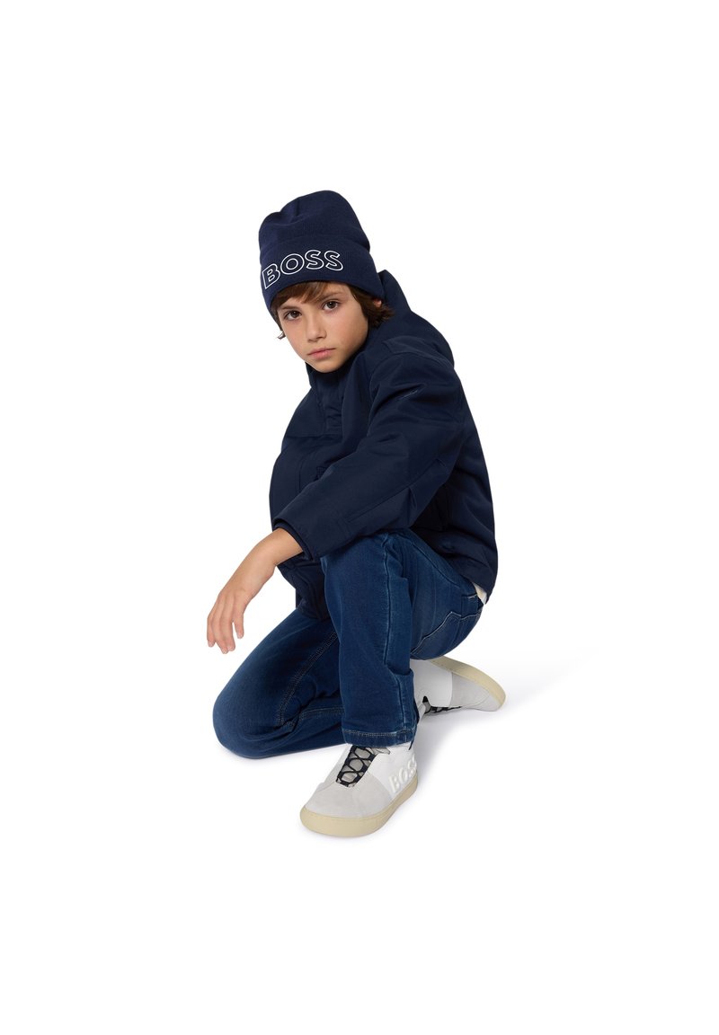 Doudoune marine, jean bleu et sneakers blancs avec des accents gris. Bonnet avec le logo "BOSS" en broderie blanche. Enfant en position accroupie.