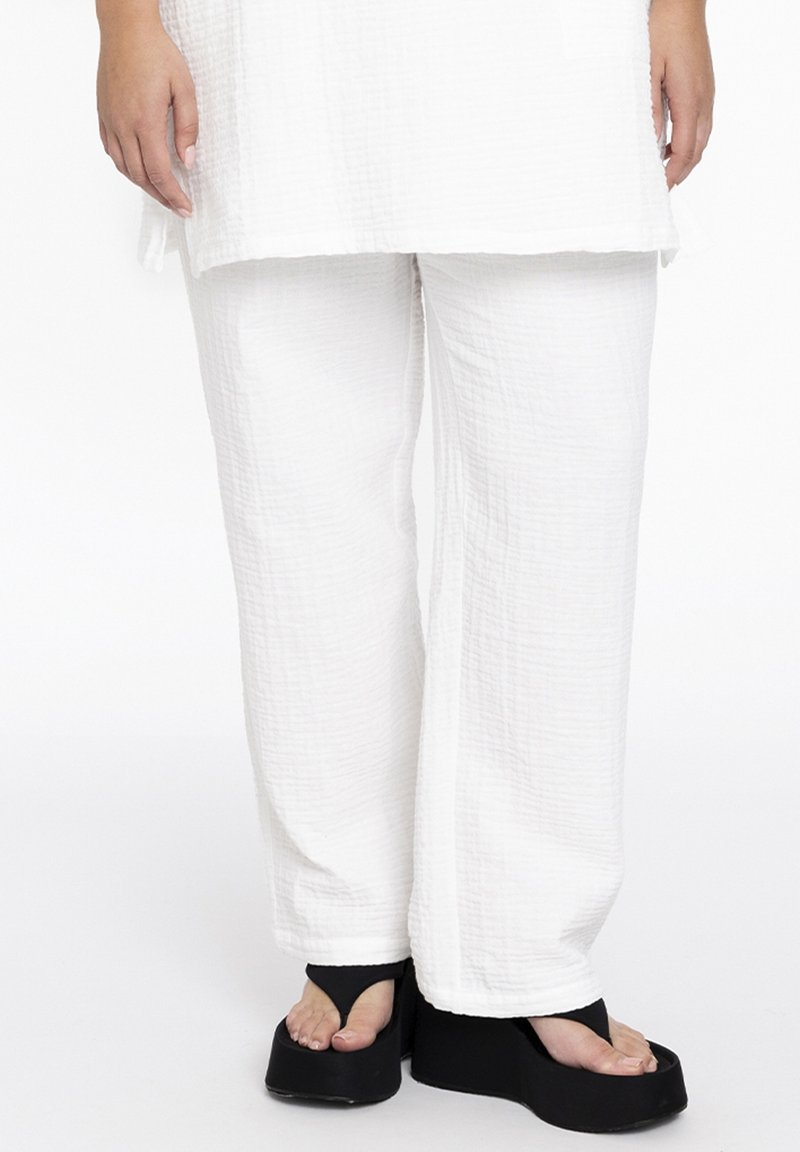 Weiße, strukturierte, locker sitzende Hosen mit einem zerknitterten Stoffmuster, kombiniert mit schwarzen Plateausandalen im T-strap-Design.