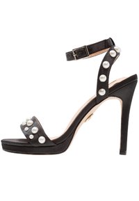 Czarne sandały na wysokim obcasie z paskiem na kostkę, ozdobione dużymi białymi perłami na paskach i na palcach.