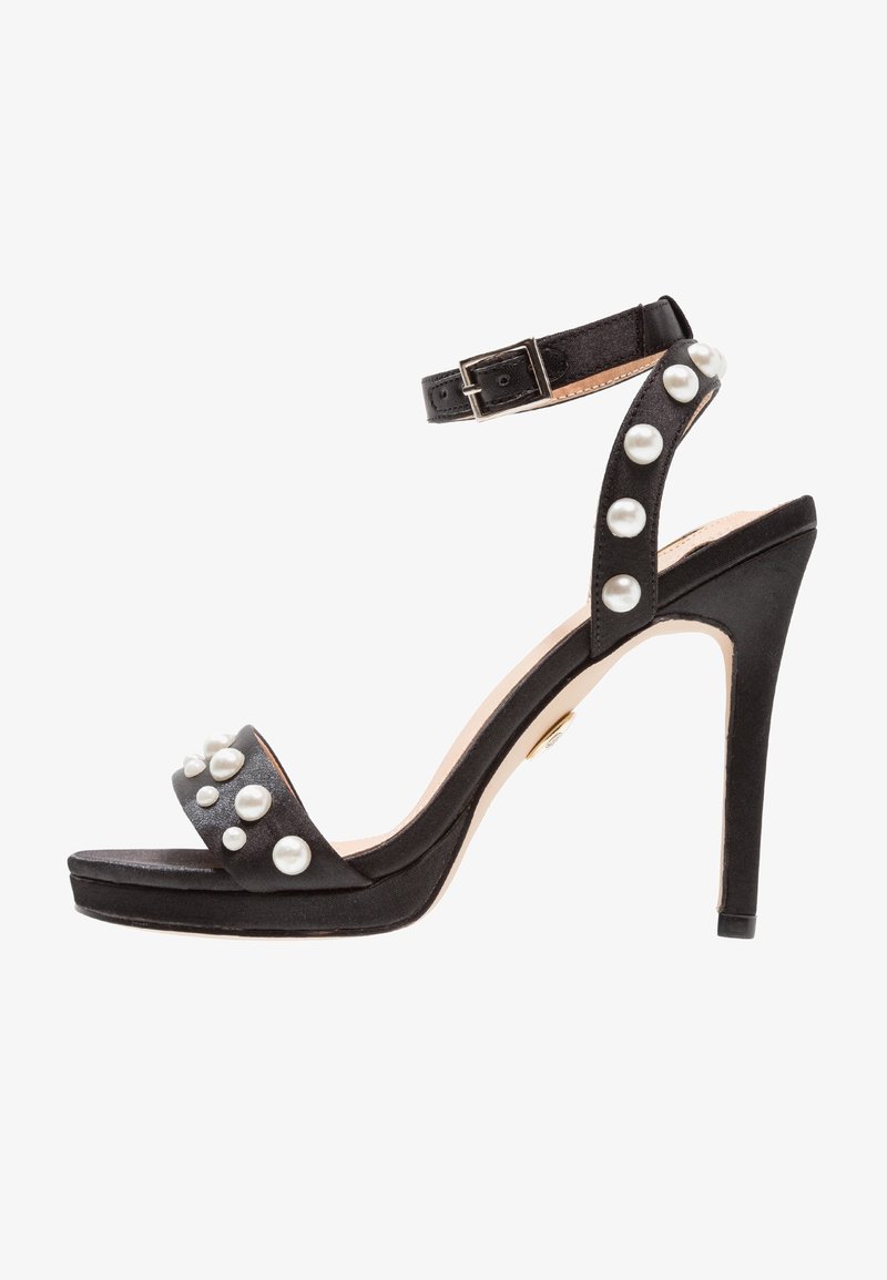 Czarne sandały na wysokim obcasie z paskiem na kostkę, ozdobione dużymi białymi perłami na paskach i na palcach.